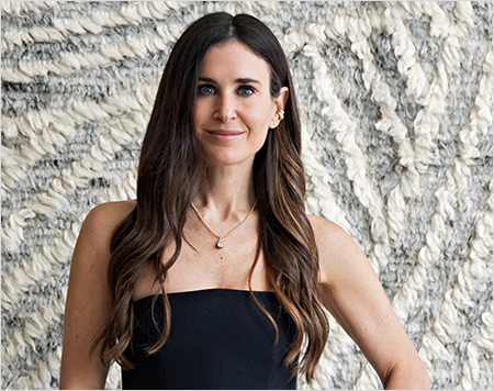 Woman with long brown hair wearing a black top against a textured stone wall.