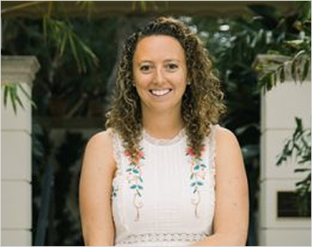 Woman with curly hair wearing a white sleeveless top with floral embroidery, standing outdoors with greenery in the background.