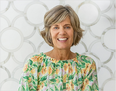 Woman with short blonde hair wearing a floral blouse against a decorative white wall.
