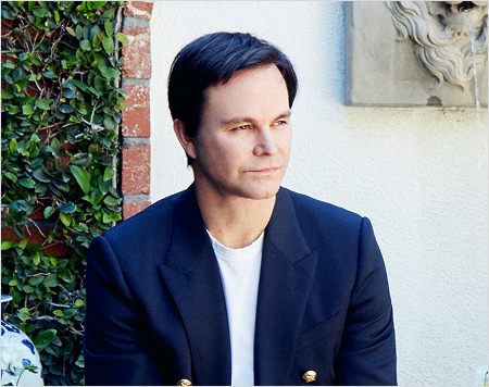 Man in a navy blazer standing outdoors with greenery and a stone wall in the background